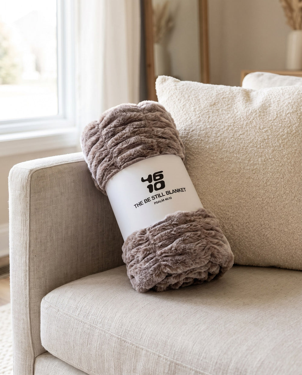 The Be Still Blanket rolled in its white paper band printed with the 46/10 logo and Psalm 46:10, on a linen sofa