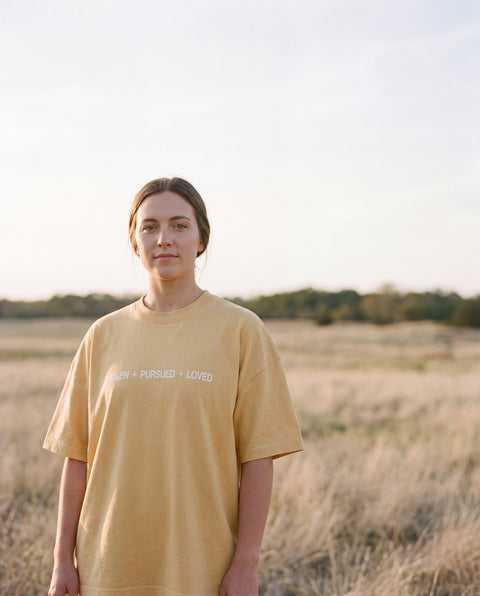Model wearing the butter yellow Chosen Pursued Loved tee in an open field at golden hour