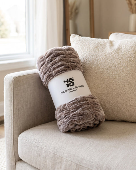 The Be Still Blanket rolled in its white paper band printed with the 46/10 logo and Psalm 46:10, on a linen sofa