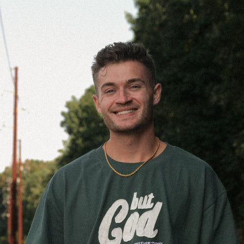 Man wearing a green t-shirt with 'but God' printed on it, standing outdoors.