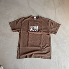 Brown oversized t-shirt with 'Come Alive' text in white, laid flat on concrete floor