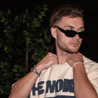 Man wearing streetwear graphic tee and black sunglasses, posing indoors with dark background.