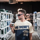 Man in sunglasses wearing streetwear tee with 'He Won't Give Up On Me' text, standing in warehouse