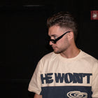 Man wearing sunglasses and a beige streetwear tee with 'HE WON'T' text, dark background