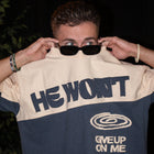 Man wearing sunglasses lifts beige and navy streetwear tee with 'He Won't Give Up On Me' text.